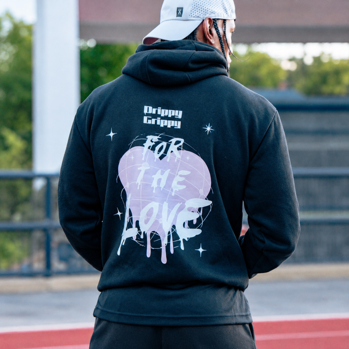 A person viewed from behind wearing a black hoodie with a large back graphic. The design features a light pink heart, white spray-paint style text reading 'FOR THE LOVE', small white stars, and the words 'Drippy Grippy' near the neck.