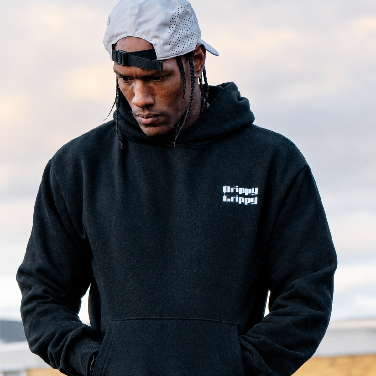 A man wearing a black hoodie with a white 'Drippy Grippy' logo on the chest, paired with a light gray mesh backward cap, photographed outdoors against a cloudy sky.