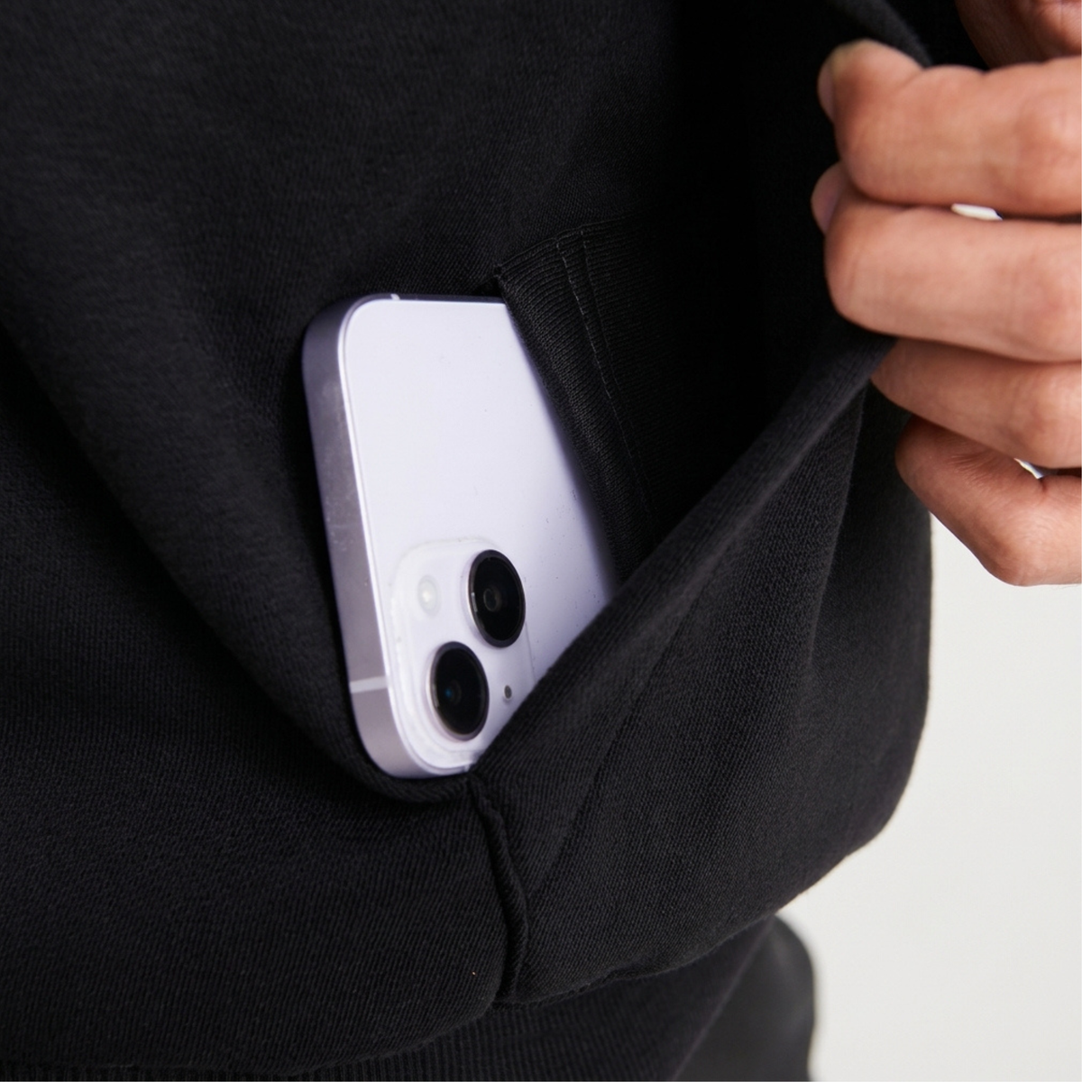A close-up of a black hoodie internal phone pocket holding a light purple smartphone, with a hand pulling the pocket open.