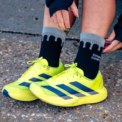 Reflective Black | A close-up of a person's legs and feet wearing bright neon yellow running shoes with dark blue stripes, paired with black and grey Drippy Grippy Endurance Grip crew socks on an asphalt surface.