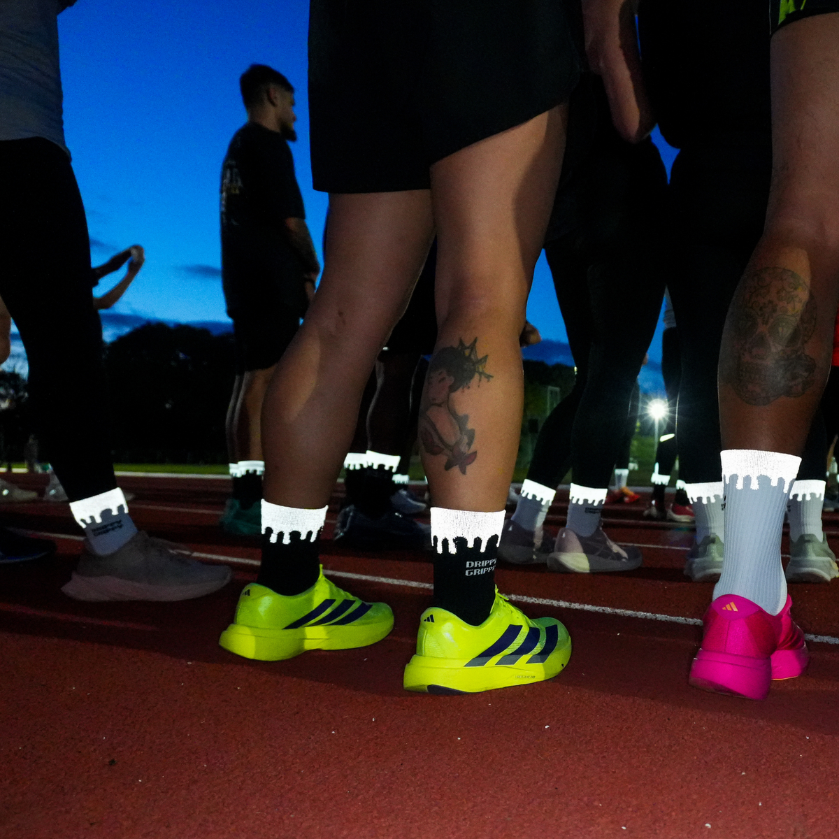 Reflective Black | A group of athletes standing on a running track at dusk, focusing on their neon running shoes and reflective socks. The central figure wears bright neon yellow-green Adidas running shoes with black socks, while others wear pink, gray, and white shoes and socks.