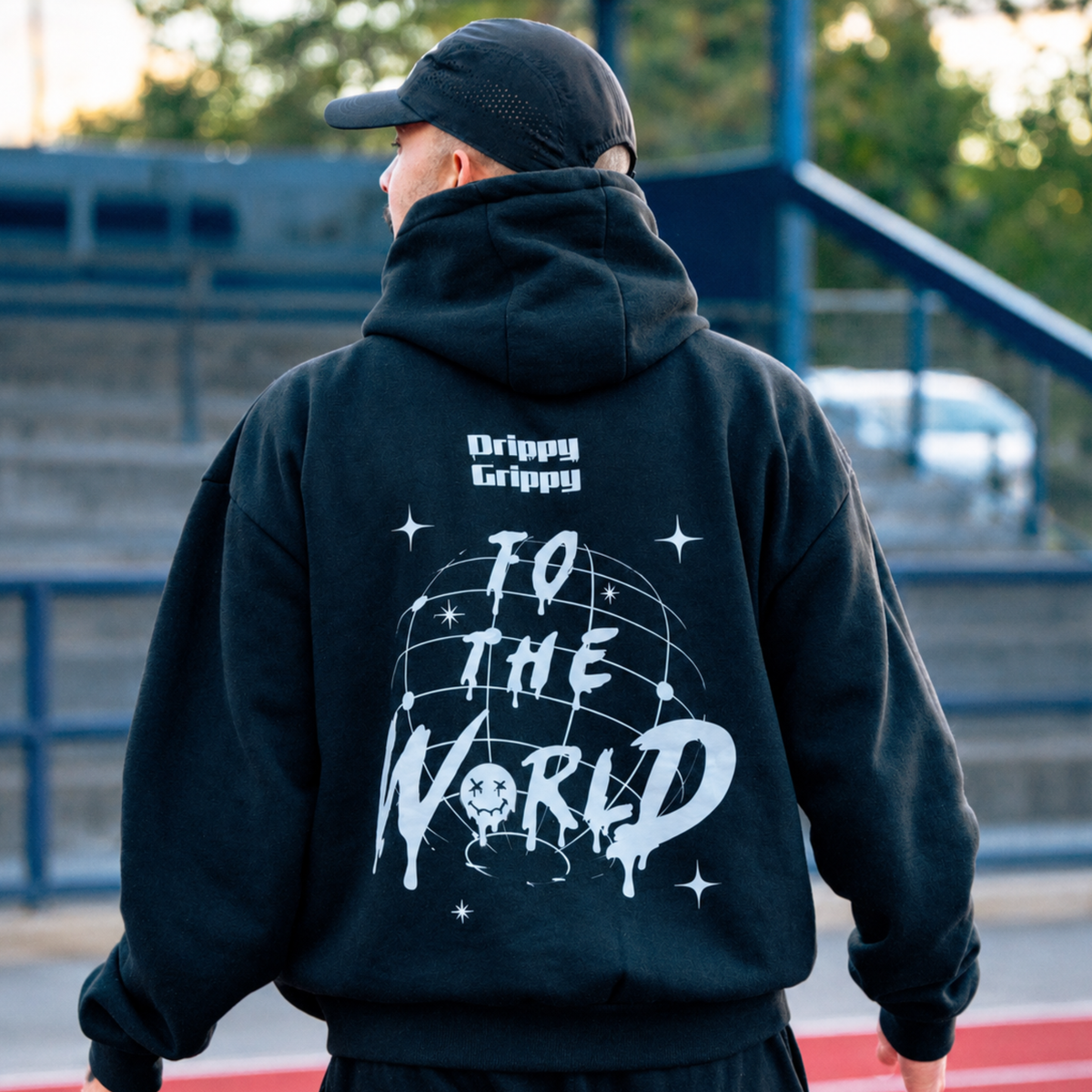 A man viewed from behind wearing a black hoodie with a large white graphic on the back that reads 'TO THE WORLD' with stars and a globe design, plus 'Drippy Grippy' text above. The setting appears to be an outdoor urban or sports field with bleachers in the background.